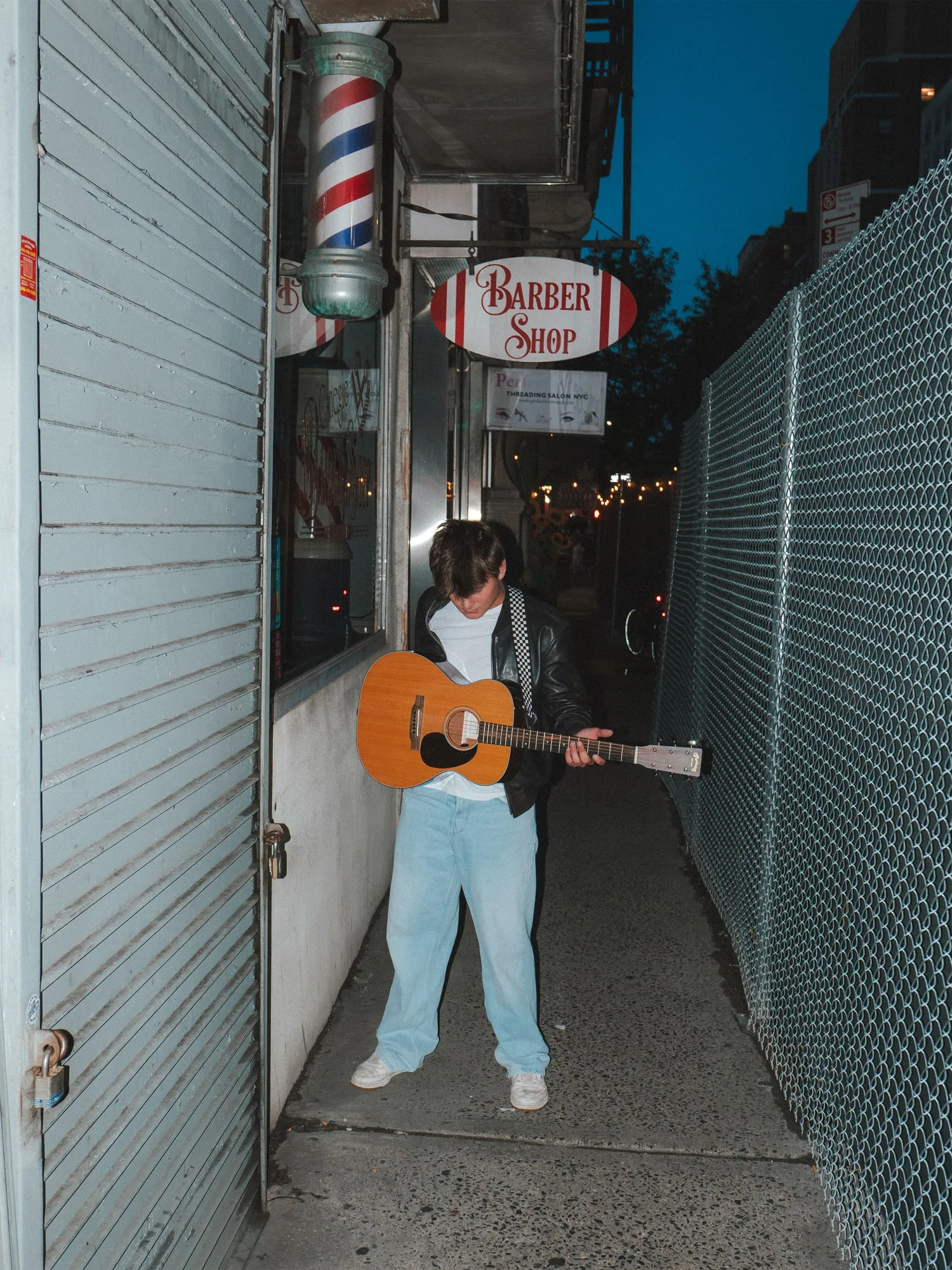 Owen Steib playing an acoustic guitar on a New York City sidewalk at dusk, under a barber shop sign